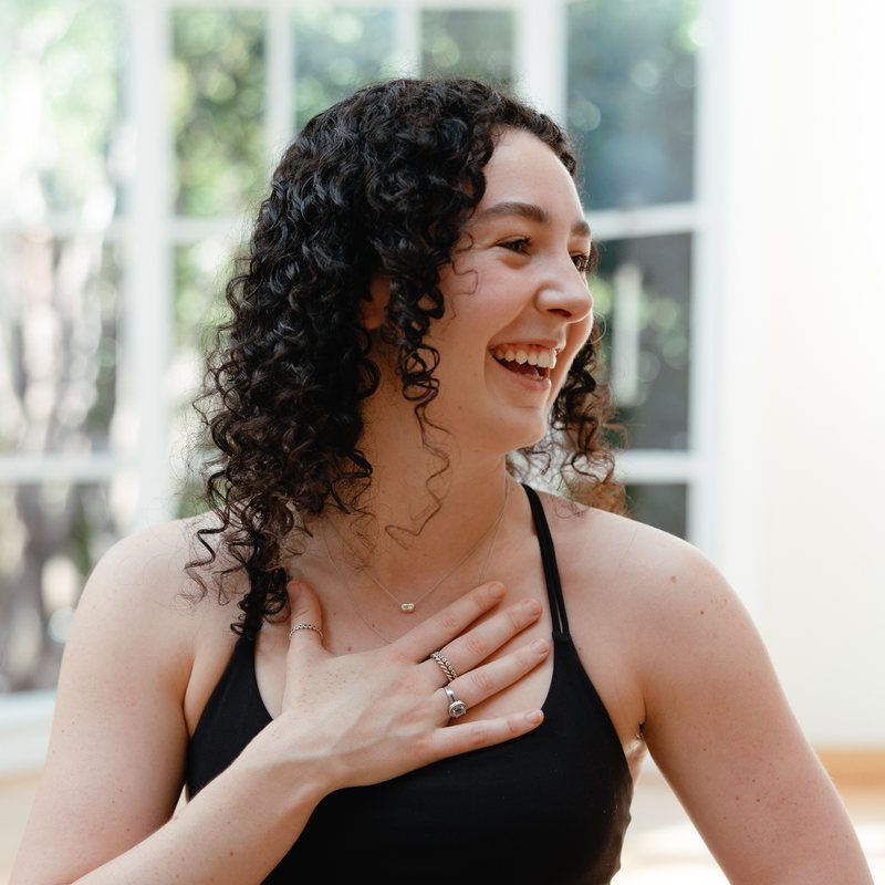 Annie Barton, vinyasa yoga teacher at The Castro Room in San Francisco, laughing with one hand on the chest, in a black top in a sunlit room with tall windows