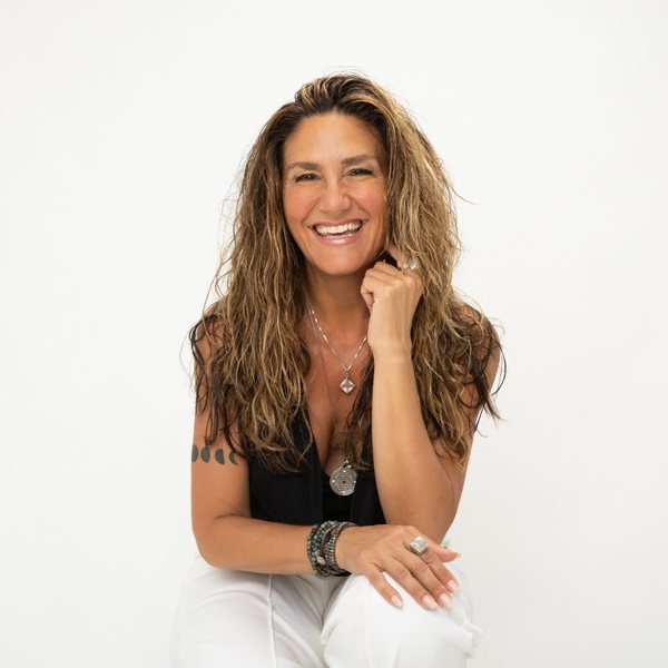Dana Damara, vinyasa yoga teacher at The Castro Room in San Francisco, smiling, with long wavy hair and layered silver jewelry, against a white backdrop