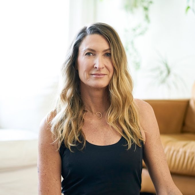 Janet Stone, vinyasa yoga teacher at The Castro Room in San Francisco, seated in a bright living room with long wavy hair, wearing a black tank top