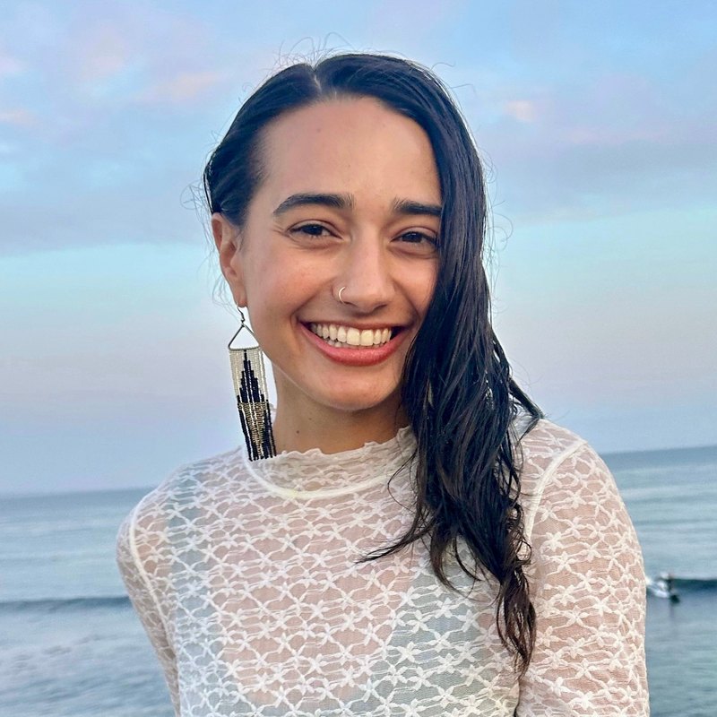 Jessica Weitzenhoffer, vinyasa yoga teacher at The Castro Room in San Francisco, smiling, with long dark hair over one shoulder and a white lace top, on an ocean bluff at dusk