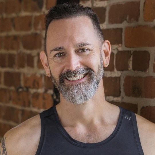Martin Scott, vinyasa yoga teacher at The Castro Room in San Francisco, smiling, with a gray beard and a black tank top, in front of a brick wall