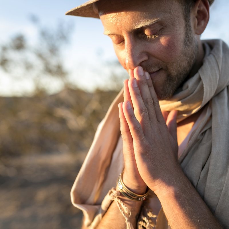 Nat Kendall, vinyasa yoga teacher at The Castro Room in San Francisco, eyes closed with hands pressed together at the lips, in a hat and scarf in desert light