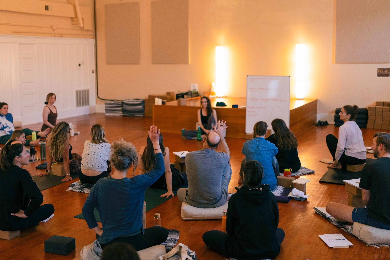 Yoga teacher training students seated on the studio floor as one raises a hand during a group discussion