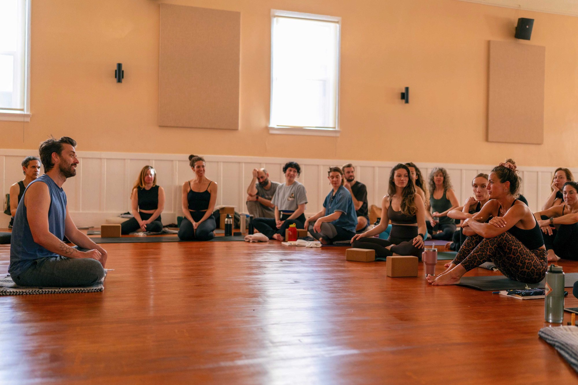 Students seated in a circle on the studio floor, smiling and listening, during yoga teacher training