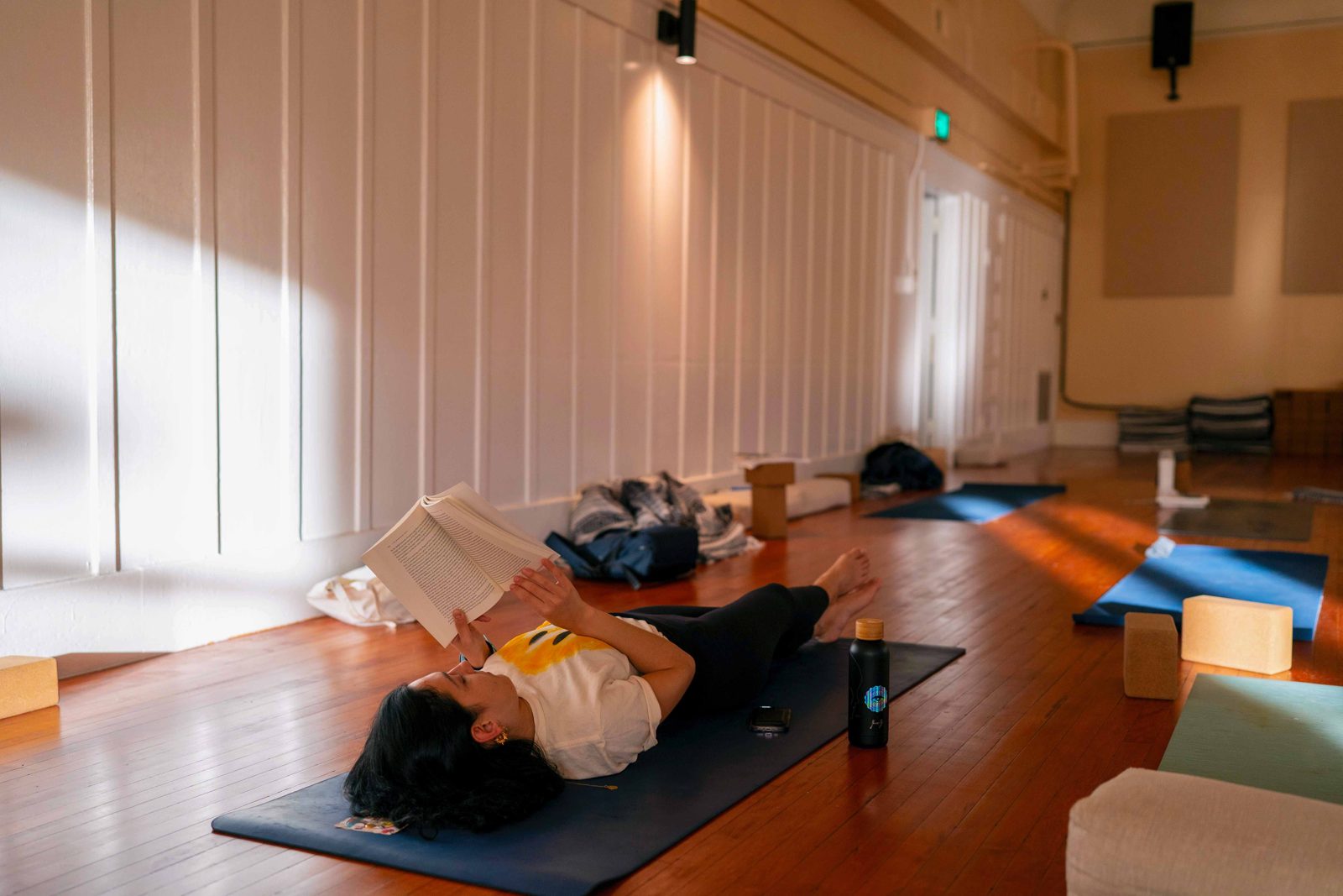 A student lies on a yoga mat reading a book in a sunlit corner of the studio