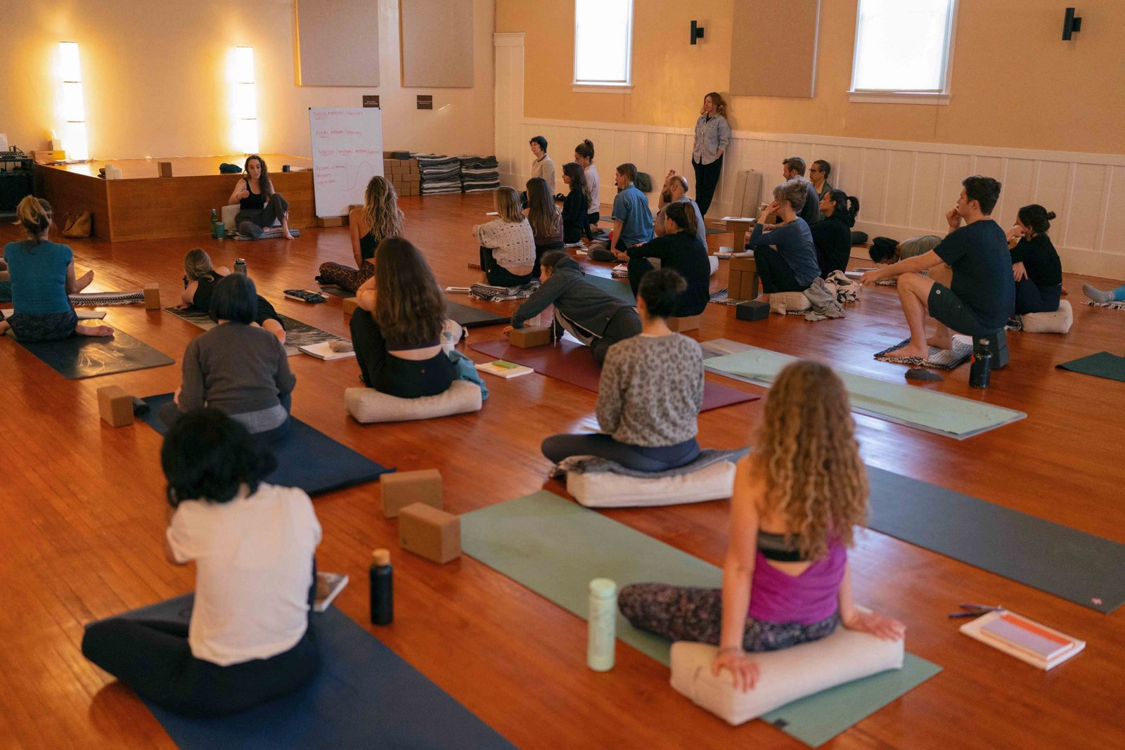 Students seated on bolsters and mats in the practice room as a teacher addresses the group